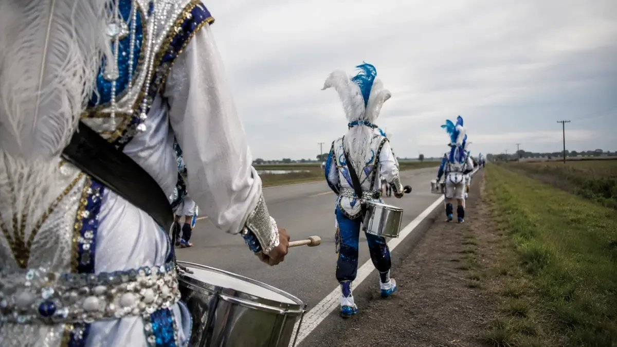 Plumas, lentejuelas y tambores sin insignias en una ruta entrerriana entre campos, plano medio documental, perspectiva natural (ilustración generada con IA)