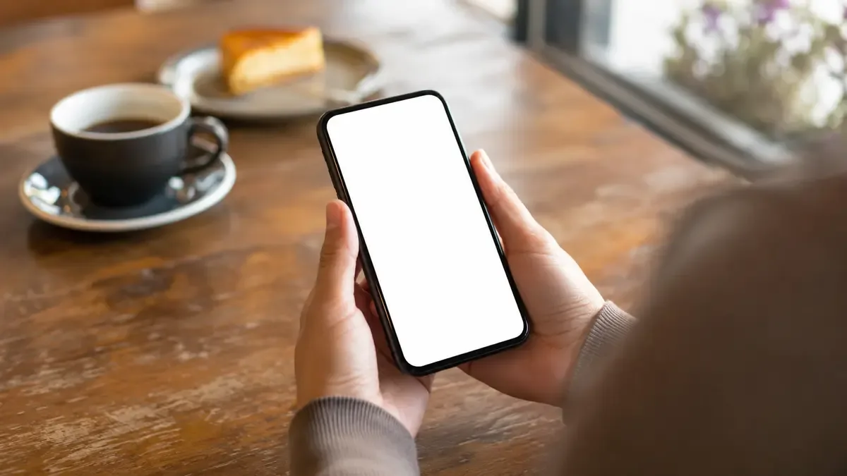 Manos sosteniendo un telefono celular de pantalla en blanco sobre una mesa de cafe (ilustración generada con IA)